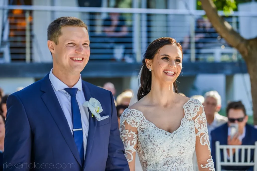 Smiling couple ceremony navy suit lace gown bokeh – Bona Dea Estate, Hermanus