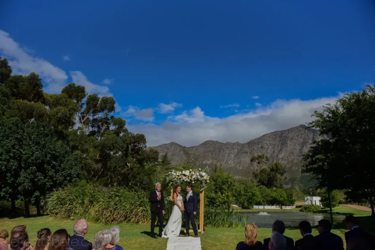 Outdoor ceremony wooden arch white flowers lakeside – Holden Manz Wine Estate, Franschhoek