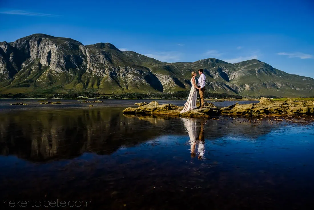 Bride groom standing rocks coastal mountain reflection – Mosaic Lagoon Café, Hermanus