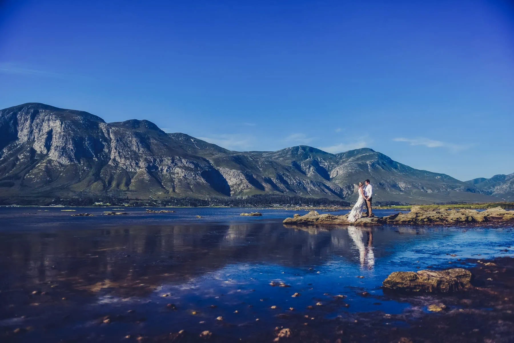Couple rocky lagoon shoreline mountain reflection – Mosaic Lagoon Café, Hermanus