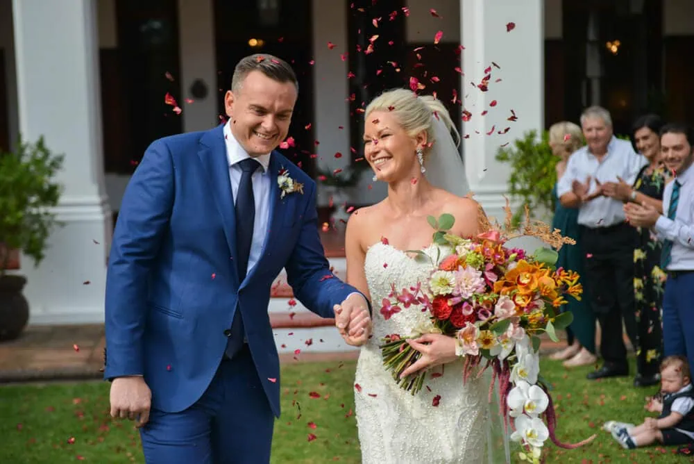 Couple rose petals white colonial building colorful bouquet – Nooitgedacht Estate, Stellenbosch